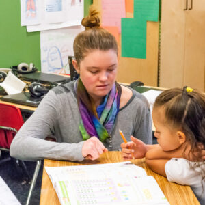Teacher helps a child with a workbook at a classroom table with colorful posters in the background.