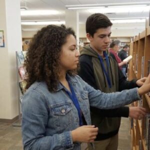Two teens browse books on a library shelf, one reaching for a title while a sign reading 'Teen Volunteers Welcome' appears on a column in the background.